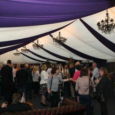A drink reception inside a marquee dressed with Brunel wall linings white ceiling linings and lots of Chesterfield sofas and black chandeliers