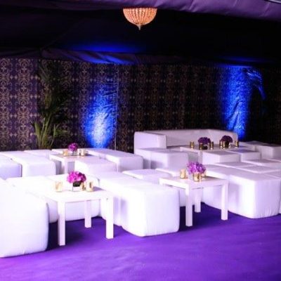 A bright white seating area with sofas cubes and coffee tableson a purple carpet inside a marquee