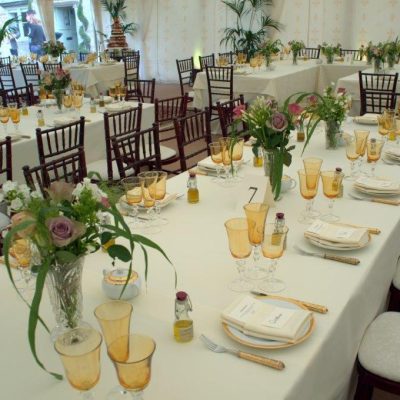 A table is set with a white table cloth golden amber glasses and some flowers inside a marquee