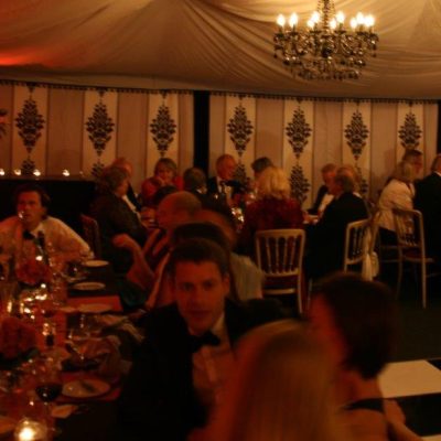 A marquee party with black white and grey linings and pink table cloths next to a chequered dance floor with guests seated and the lights on