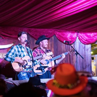 A group of musicians dressed as cowboys sing in a band standing on a marquee stage in front of themed marquee linings that look like real wood plank walls